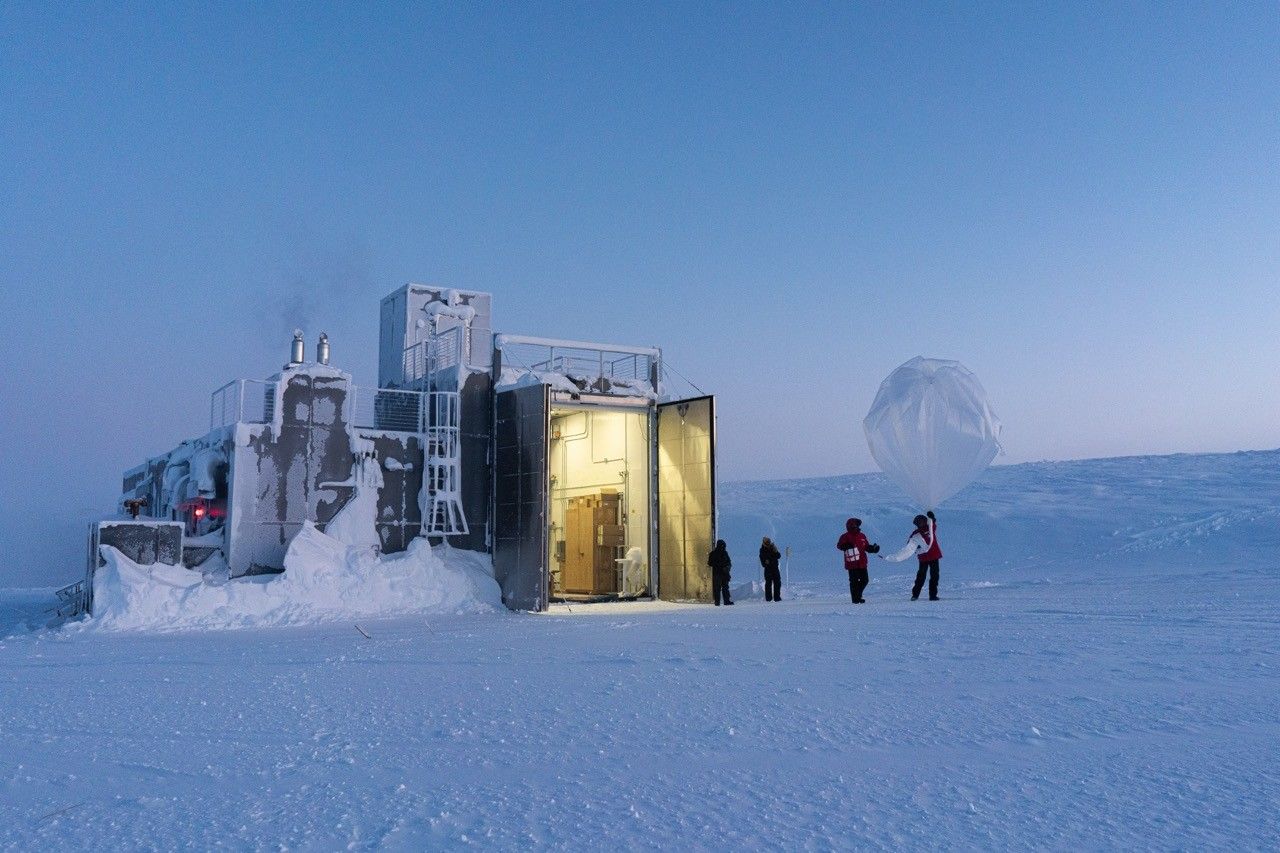 a partially snow encased metal building with a large open door to a utility space sits in the background of wholly snow covered landscape with two people holding onto a translucent plastic looking balloon which is roughly the diameter of the person holding it.